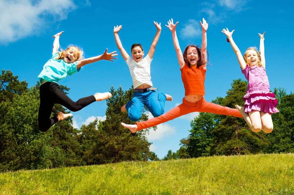 Vier Kinder springen mit gegrätschten Beinen und Händen auf einer Wiese in die Höhe.